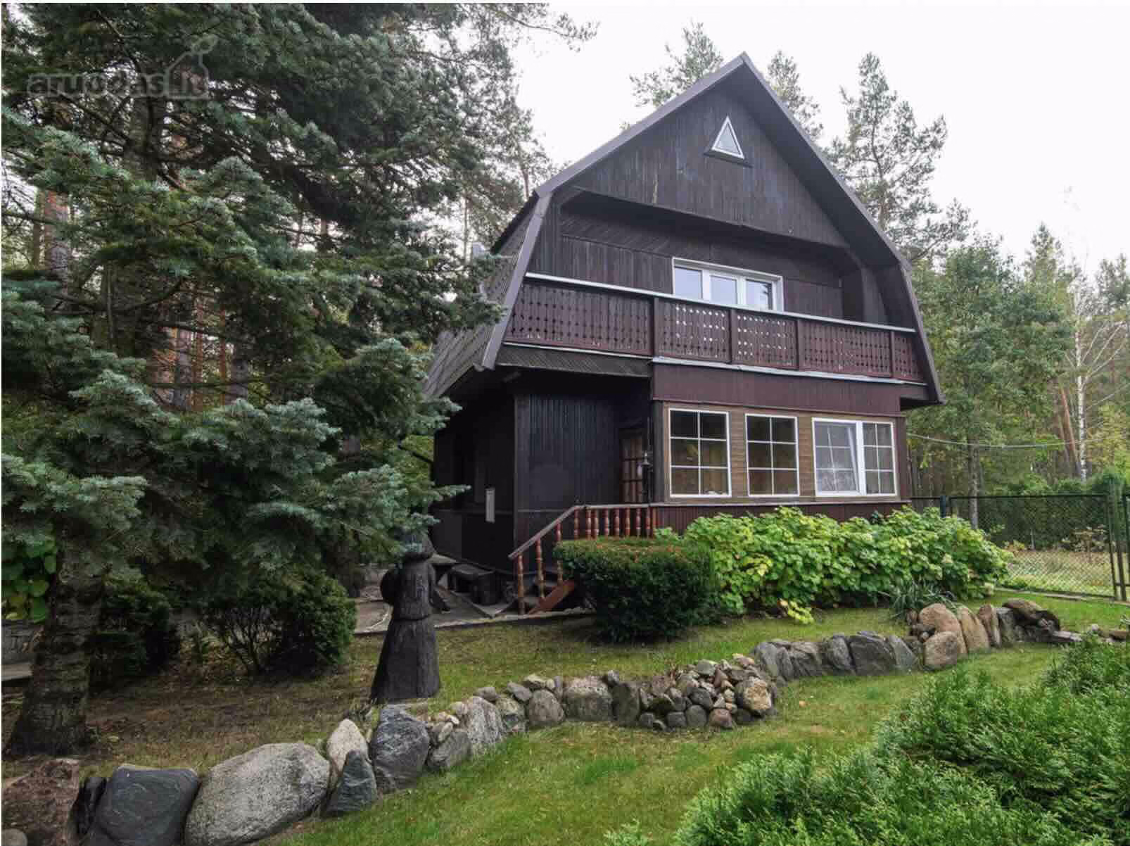 Two-story dark wooden house with balcony, surrounded by pine trees and stone landscaping.