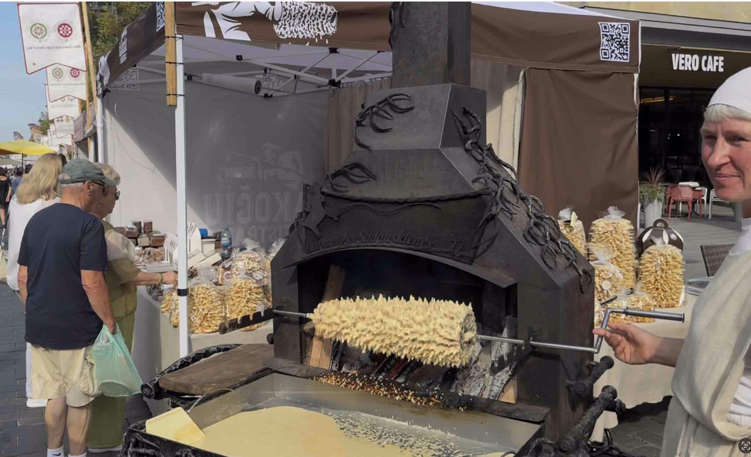 A large, ornate metal oven is shown with a spit cake, a traditional pastry, baking over an open fire.